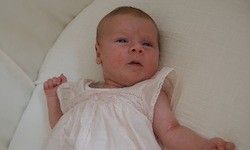 Ein kleines Baby, kahl und in einem weißen Kleid, liegt auf einem weißen Kissen und scheint etwas anzuschauen.