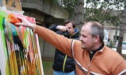 Ein Mann in einer orangefarbenen Jacke zeigt auf ein lebhaftes Gemälde, während ein anderer Mann in einer schwarzen Jacke hinter ihm beobachtet.