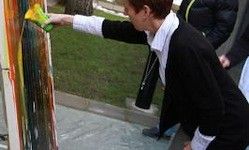 Eine Person malt ein buntes abstraktes Wandbild an einer Wand mit einer grünen Sprühdose. Eine andere Person steht hinter ihr.