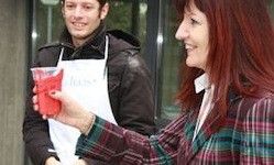 Eine Frau mit roten Haaren hält einen roten Becher und lächelt, während ein Mann in einer weißen Schürze und einem schwarzen Mantel sie ansieht.