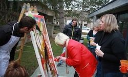 Eine Gruppe von Menschen, darunter zwei Frauen und ein Mann, malen auf Leinwänden vor einem Gebäude. Die Frau in Rot vervollständigt ihr Bild, während die anderen beobachten.