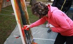 Eine Frau in einem rosa Hoodie malt ein großes Gemälde mit mehreren Farben auf einem Grasfeld. Sie hält einen Pinsel und scheint konzentriert zu arbeiten.