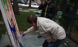 Ein Mann malt eine große Leinwand mit blauer Farbe. Er beugt sich vor und hält einen Pinsel. Ein anderer Mann steht hinter ihm und macht ein Foto. Ein Auto ist am Straßenrand geparkt.