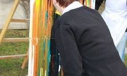 Eine Person in einer schwarzen Jacke betrachtet ein farbenfrohes Gemälde. Die Leinwand ist auf einem hölzernen Staffelei mit einer dreieckigen Struktur montiert. Eine weitere Person in einer weißen Schürze steht in der Nähe.