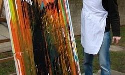 Eine Person in einer weißen Schürze und Jeans steht neben einem farbenfrohen abstrakten Gemälde auf einem Staffelei. Der Hintergrund ist ein Grasbereich mit einem Steinweg.