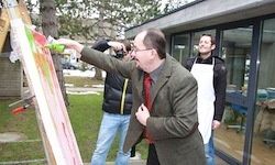 Ein Mann in Anzug und Krawatte malt im Freien auf eine große Leinwand. Zwei andere Männer in Schürzen stehen in der Nähe, einer hält eine Kamera.