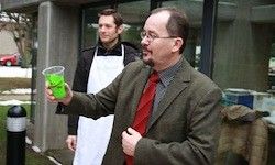 Ein Mann in Anzug und roter Krawatte hält einen grünen Drink vor einem Gebäude, hinter ihm steht ein weiterer Mann in einer weißen Schürze.