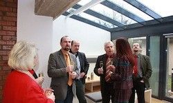 Eine Gruppe von Menschen in formeller Kleidung steht und hält Weingläser in einem Raum mit Glasdach. Der Raum hat Ziegelwände und Glastüren. Sie scheinen ein Gespräch zu führen.