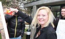 Eine Frau mit blondem Haar steht draußen, hält eine Leiter und lächelt. Sie scheint etwas zu reparieren. Hinter ihr stehen zwei Männer, einer trägt ein weißes Küchentuch. Bäume sind im Hintergrund zu sehen.