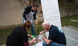 Drei Personen arbeiten zusammen im Freien, eine gießt Flüssigkeit ein, eine andere hält eine Flasche und die dritte steht in der Nähe einer großen weißen Plane.