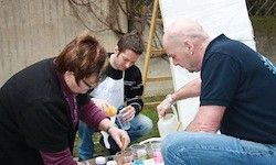 Drei Personen arbeiten gemeinsam im Garten. Eine Frau rührt etwas in einer Schüssel, während eine andere Person eine Flüssigkeit hinzufügt. Die dritte Person hält einen Messbecher.