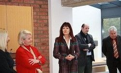 Fünf Erwachsene stehen in einem Raum, möglicherweise bei einer Besprechung. Eine Frau in einem roten Mantel und eine andere in einem karierten Mantel haben die Arme verschränkt. Ein Mann mit Krawatte steht mit verschränkten Armen.