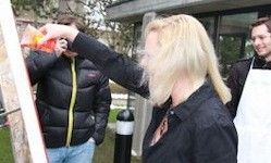 Eine Frau mit blonden Haaren hält eine Flasche in der Hand und trägt wahrscheinlich etwas auf eine Wand auf. Ein Mann in einem schwarzen Jackett steht hinter ihr, und ein weiterer Mann in einer weißen Schürze ist in der Nähe.
