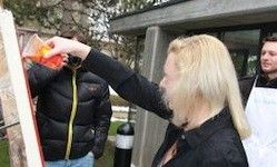 Eine Frau mit blonden Haaren malt mit orangefarbener Farbe auf eine Leinwand. Ein Mann in einer schwarzen Jacke steht hinter ihr. Ein weiterer Mann in einer weißen Schürze steht an der Seite. Bäume und ein Gebäude sind im Hintergrund.