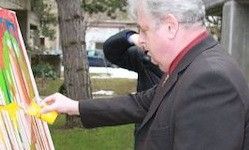 Ein älterer Mann in einem schwarzen Anzug steht im Freien und malt eine farbenfrohe Leinwand auf einem Staffelei. Er hält einen Pinsel in der rechten Hand, und ein gelber Farbklecks ist auf seiner linken. Eine Person steht hinter ihm, teilweise sichtbar.