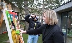 Eine Frau in einem schwarzen Mantel steht draußen und malt auf eine Leinwand mit einem Pinsel. Ein Mann mit einer Kamera hinter ihr beobachtet. Bäume und ein Gebäude sind im Hintergrund.
