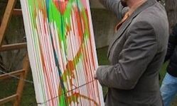 Ein Mann in einem grauen Anzug hält ein farbenfrohes abstraktes Gemälde, während er draußen steht. Das Gemälde befindet sich auf einem hölzernen Staffelei. Ein weiterer Mensch ist im Hintergrund teilweise sichtbar.