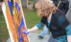 Eine Frau malt ein großes Leinwand mit blauen und orangefarbenen Farben im Freien. Eine andere Person steht hinter ihr auf dem Gras.
