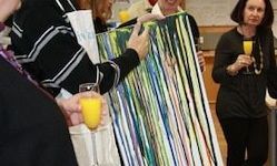 Eine Gruppe von Menschen hält Gläser mit Orangensaft und einem bunten Gemälde, wahrscheinlich in einer Küche. Eine Person hält das Gemälde, während andere zusehen.