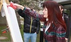 Eine Frau mit roten Haaren hält ein Werkzeug und scheint an einer großen weißen Tafel zu arbeiten. Sie trägt eine karierte Jacke. Hinter ihr steht ein Mann in einer schwarzen Jacke, der zusieht.