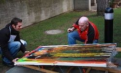 Zwei Männer arbeiten an einem großen, farbenfrohen Gemälde im Freien. Ein Mann kniet und trägt eine schwarze Jacke, während der andere gebückt ist und eine rot-schwarze Jacke trägt. Sie befinden sich auf einem Rasen mit einer Wand und einem schwarzen Pfosten im Hintergrund.