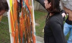 Eine Frau in einem schwarzen Shirt und mit einer Halskette steht im Freien und malt auf eine abstrakte Leinwand mit kräftigen orangen und blauen Strichen. Hinter ihr beobachtet eine Person in Jeans.