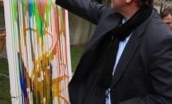 Ein Mann in einem schwarzen Anzug und Schal steht vor einem großen abstrakten Gemälde mit leuchtenden Farben. Er scheint das Gemälde genau zu betrachten.