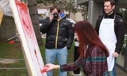 Drei Personen stehen vor einer großen Leinwand mit roten Farbspritzern. Eine Frau in einem karierten Shirt hält einen roten Becher. Ein Mann macht ein Foto mit einer Kamera. Ein anderer Mann trägt eine weiße Schürze.