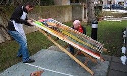 Zwei Männer arbeiten draußen an einem großen, bunten Gemälde auf einem geneigten Brett. Einer steht, der andere sitzt auf dem Boden. Ein Baum und ein Pfosten sind in der Nähe, und Schnee liegt auf dem Boden.