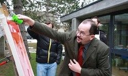 Ein Mann sprüht rote Farbe auf eine Leinwand auf einem Staffelei im Freien. Er trägt einen Anzug und eine Brille, während ein anderer Mann in einem Mantel hinter ihm steht. Bäume und ein Gebäude sind im Hintergrund.