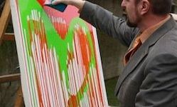 Ein Mann in einem grauen Anzug malt ein leuchtendes, abstraktes Herz auf eine große Leinwand. Das Gemälde zeigt kräftige Striche in Grün, Orange und Pink.