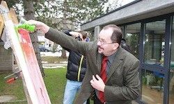 Ein Mann in Anzug und Krawatte malt draußen auf einer großen Leinwand. Ein Mensch mit einer Kamera steht hinter ihm, und ein Gebäude mit Glasfenstern ist im Hintergrund.