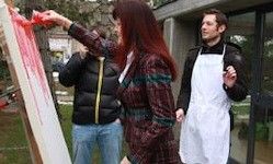 Eine Frau mit roten Haaren malt auf eine Leinwand vor einem modernen Gebäude. Ein Mann in einer weißen Schürze und eine weitere Person in einer schwarzen Jacke stehen in der Nähe.
