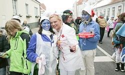 Zwei Frauen in Kostümen, eine mit blauem Gesicht und die andere in Weiß, stehen zusammen auf einer Straße mit mehreren Leuten, darunter ein Mann mit einem roten Kreuz auf seinem Shirt.