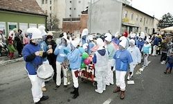 Eine Gruppe von Menschen in blauen und weißen Kostümen und weißen Mützen zieht auf der Straße, spielt Trommeln und trägt Luftballons.