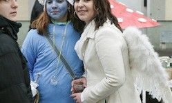 Drei Personen in Kostümen stehen draußen; eine in einem Engel-Outfit, eine andere in einem blauen Kostüm und eine dritte in Schwarz. Sie scheinen für ein Foto zu posieren.