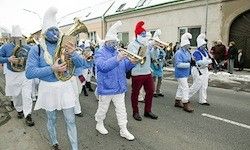 Eine Gruppe von Musikern in blauen Kostümen und weißem Gesichtsmake-up spielt Blechblasinstrumente bei einem Umzug auf einer verschneiten Straße.