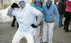 Drei Menschen mit blauer Gesichtsbemalung und passenden Outfits stehen auf einer verschneiten Straße, einer hält eine Messing-Trompete und ein Saxophon. Hinter ihnen laufen mehrere Menschen.