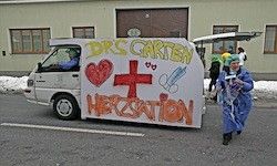 Ein weißer Lieferwagen mit Herz, Kreuz und medizinischen Symbolen auf der Seite. Eine Person in blauer Kleidung geht neben ihm und hält ein Buch. Der Lieferwagen trägt 'DRS CARTEN' und 'HYPERESTION'. Ein Gebäude mit Fenstern ist im Hintergrund.