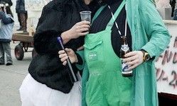 Zwei Frauen in Schürzen, eine in Grün und die andere in Schwarz, stehen zusammen und halten eine Flasche und ein Glas, mit einem unscharfen Hintergrund von Menschen und einem Karren.