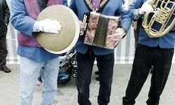 Drei Musiker in blauen Uniformen, einer hält eine Trommel, einer ein Akkordeon und einer eine Tuba, stehen nebeneinander.
