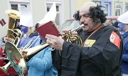 Ein Mann in Schwarz spielt Trompete und hält ein Buch in der Hand. Andere Menschen spielen hinter ihm Blechblasinstrumente.