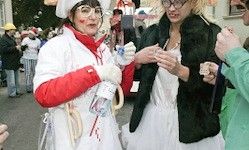 Zwei Frauen in Kostümen, eine als Krankenschwester mit Blut an den Handschuhen und die andere in einem schwarzen Mantel, stehen auf einer Straße mit Menschen und Gebäuden im Hintergrund.