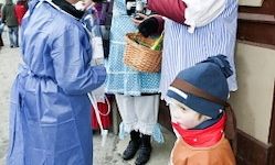 Ein junger Junge in einem blauen Hut und einer orangefarbenen Jacke schaut auf eine Person in traditioneller Kleidung, die einen Korb hält. Dahinter steht eine weitere Person in einem blauen Mantel, und andere sind im Hintergrund zu sehen.