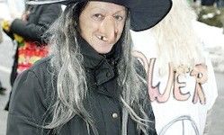 Eine Person mit langen grauen Haaren, die einen schwarzen Hut und Mantel trägt, steht auf einer Stadtstraße. Dahinter trägt eine verschwommene Figur ein weißes Shirt mit dem Wort 'WER' darauf.