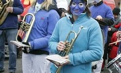 Eine Frau mit blauer Gesichtsbemalung hält eine Trompete und steht neben anderen, die Musikinstrumente halten, möglicherweise bei einer Parade. Sie tragen alle blaue Jacken und weiße Hosen.
