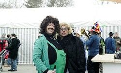 Zwei Menschen in Kostümen stehen nebeneinander bei einer Veranstaltung im Freien. Ein Mann hält eine Flasche, und eine Frau steht neben ihm. Im Hintergrund tummeln sich Menschen.