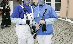 Zwei Personen mit blauem Gesichts-Make-up stehen auf einem gepflasterten Weg und halten Klarinetten. Hinter ihnen steht eine verschwommene Figur.