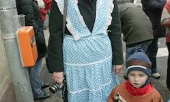 Eine Frau in einem gepunkteten Kleid und ein Kind in einem Schal gehen auf dem Bürgersteig, mit Leuten im Hintergrund.