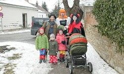 Eine Familie in Kostümen steht im Schnee, mit einem Kinderwagen in der Nähe. Sie sind auf einer Straße mit einem Auto und einem Haus im Hintergrund.
